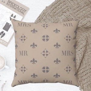 Puede incluir: Una almohada beige con un patr&oacute;n repetido de flores de lis y una cruz. La almohada tiene las palabras "MR" y "MRS" impresas en ella en una fuente gris.