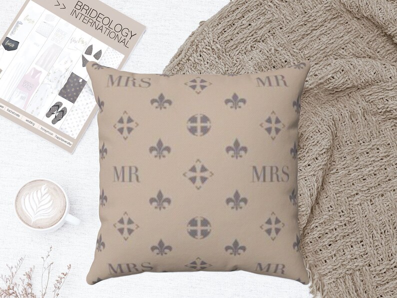 Puede incluir: Una almohada beige con un patr&oacute;n repetido de flores de lis y una cruz. La almohada tiene las palabras "MR" y "MRS" impresas en ella en una fuente gris.