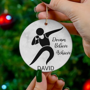 May include: White ceramic Christmas ornament with a black silhouette of a person throwing a shot put. The ornament has the text "Dream Believe Achieve" and the name "DAVID" printed on it.
