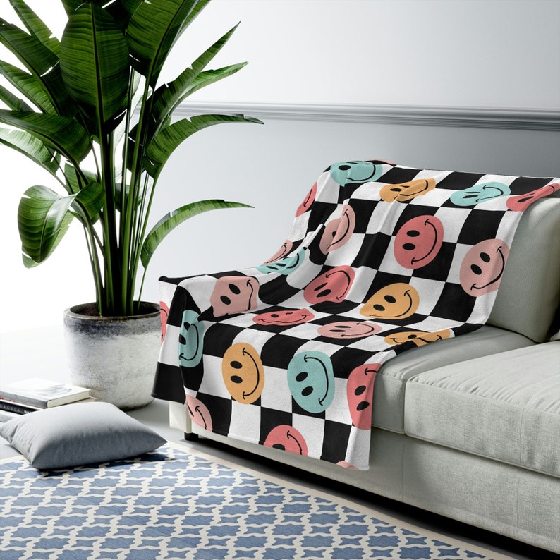 May include: A white throw blanket with a black and white checkered pattern and colorful smiley faces. The blanket is draped over a light gray couch.