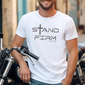 Pu&ograve; includere: Maglietta bianca con una croce grigia e il testo "STAND FIRM 1 Cor 16:13".