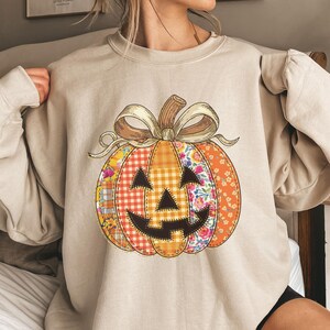 May include: Beige sweatshirt featuring a patchwork pumpkin design. The pumpkin has a smiling jack-o'-lantern face and is adorned with floral, plaid, and orange patterns. A beige bow sits atop the pumpkin. Perfect for fall and Halloween.