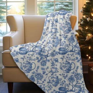 May include: A white throw blanket with a blue and white floral pattern featuring teacups and teapots. The pattern is reminiscent of vintage china.