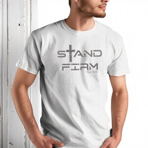 Pu&ograve; includere: Una maglietta bianca con una grafica grigia che dice "STAND FIRM" con una croce sopra la parola "STAND" e il testo "1 Cor 16:13" sotto la parola "FIRM".