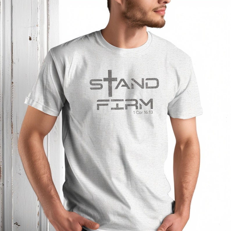 Pu&ograve; includere: Una maglietta bianca con una grafica grigia che dice "STAND FIRM" con una croce sopra la parola "STAND" e il testo "1 Cor 16:13" sotto la parola "FIRM".