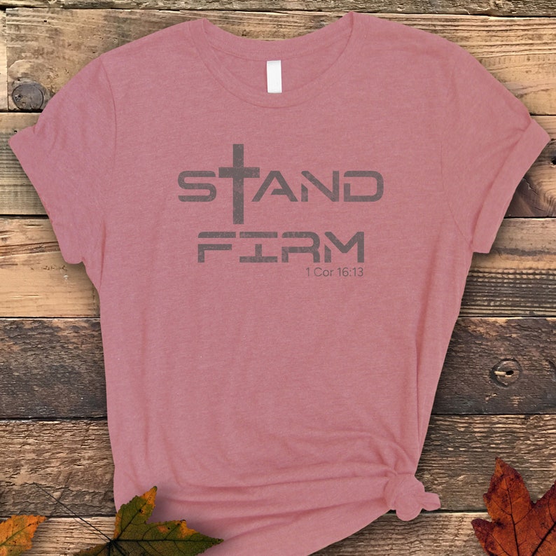 Pu&ograve; includere: Una maglietta rosa chiaro melange con una grafica grigia che dice "Stand Firm" con una croce sopra la parola "Stand" e il riferimento biblico "1 Cor 16:13" sotto la parola "Firm".