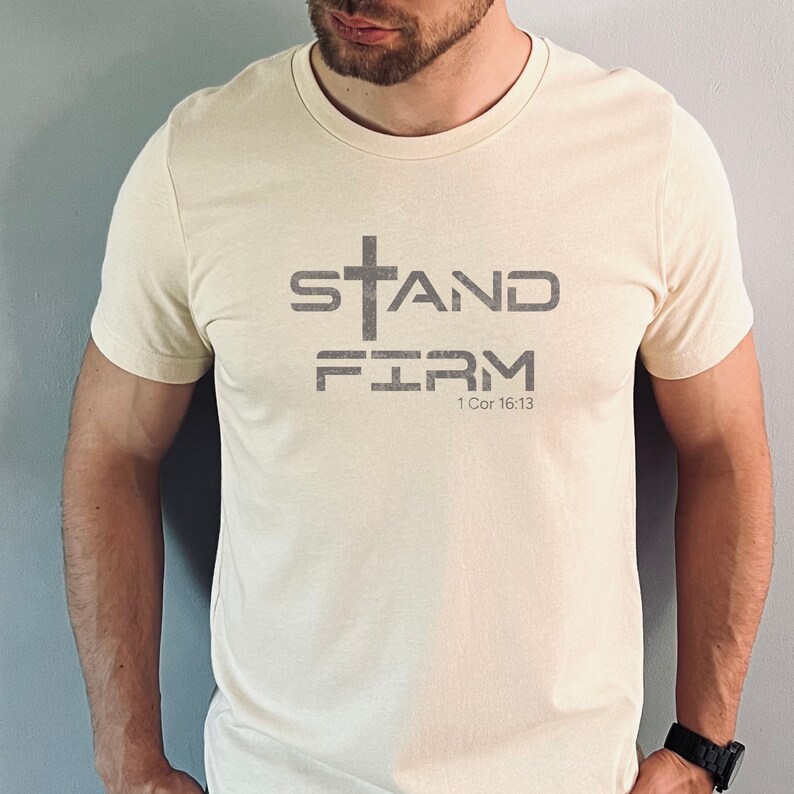 Pu&ograve; includere: Una t-shirt color crema con una grafica grigia che dice "STAND FIRM" con una croce sopra la parola "STAND" e il testo "1 Cor 16:13" sotto le parole.
