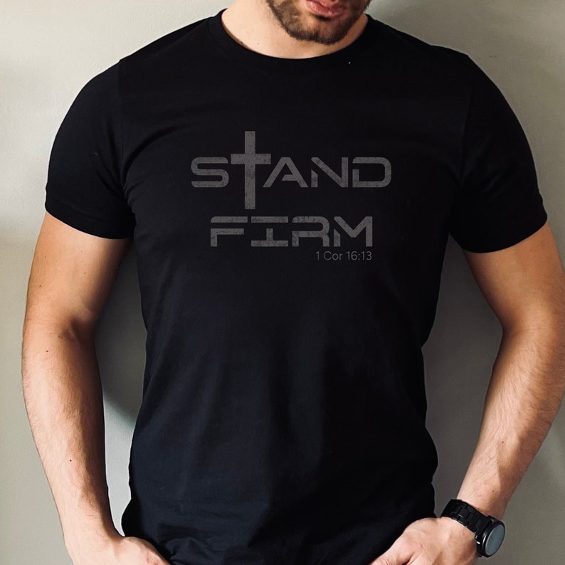 Pu&ograve; includere: Maglietta nera con una croce bianca e il testo "STAND FIRM" e "1 Cor 16:13".