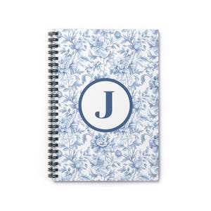 May include: A blue and white spiral notebook with a floral pattern and a circle with the letter "J" in the center.