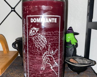 May include: A burgundy candle in a clear glass jar. The label reads "DOMINANTE" and "DOMINATION" with a white graphic of an eye, lightning, and figures. The candle is on a shelf with other decorative items.