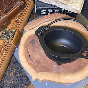 Puede incluir: Un pequeño caldero de hierro fundido negro con asa, sobre una rodaja de madera. En el fondo se ven dos diarios encuadernados en cuero con cierres metálicos. El caldero tiene tres patas pequeñas y está diseñado para uso ritual.