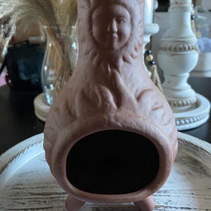 May include: A terracotta-colored decorative chiminea with a detailed face design. The chiminea has a large opening and rests on three small feet. The background includes a white tray and decorative elements.