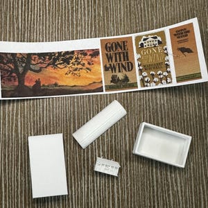May include: Miniature paper book covers with the title "Gone With The Wind" and a sunset scene. Also, white miniature accessories, including a rectangular box, a rolled-up item, a small architectural piece, and a rectangular tray.