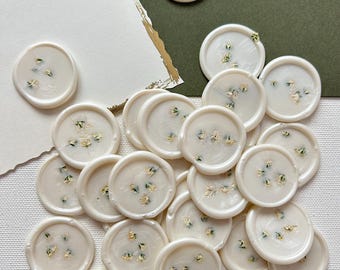 Puede incluir: Una colección de 20 sellos de cera blanca con flores secas incrustadas en la cera. Los sellos son redondos y tienen un borde ligeramente elevado. Las flores son de color verde claro y blanco.
