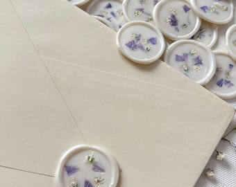 Puede incluir: Primer plano de un sobre blanco con un sello de cera blanco con flores secas de color púrpura y blanco. El sello está en la esquina superior derecha del sobre.