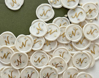 Puede incluir: Una colección de sellos de cera blanca con una letra dorada "N" y pequeñas flores blancas incrustadas en la cera.