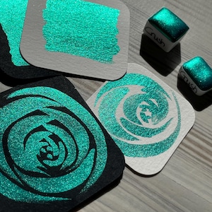 May include: Four samples of teal green glitter paint on white and black paper squares. Two small teal green glitter paint pans labelled "Crush" are also shown.