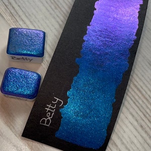 May include: Two small square jars of blue and purple iridescent glitter paint with the word "Betty" written on the labels. The paint is shown on a black card with a gradient of blue and purple glitter paint.