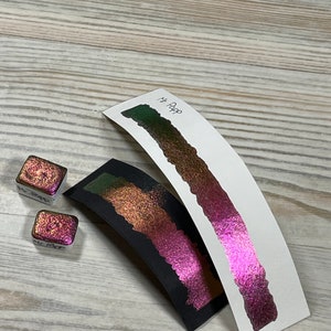 May include: Two small square containers of iridescent pink and green paint with a sample of the pink paint on a white card labeled "Mt. Pop".