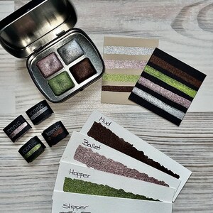 May include: A set of four eyeshadows in a silver tin with a black lid. The eyeshadows are in shades of green, brown, pink, and silver. The eyeshadows are labeled "Iconic" and the swatches are labeled "Mud", "Ballet", "Hopper", and "Slipper".