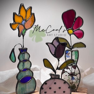May include: A stained glass pattern for a floral trio, featuring three vases with flowers in shades of pink, purple, orange, and red. The vases are on a wooden base. The pattern is titled "Floral Trio Set 1" and is by McCool's Art Studio.