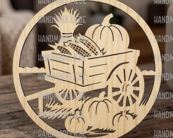 May include: A circular light-colored wooden cutout featuring a laser-cut design of a wagon filled with corn and a pumpkin, with additional pumpkins resting on the ground below. The piece includes a small hole at the top for hanging purposes.