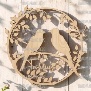 Pode incluir: Uma decoração de parede circular em madeira clara com um design cortado a laser de dois pássaros pousados num ramo dentro de uma coroa de folhas. A palavra Fruehling está escrita em letra cursiva no centro inferior do recorte.