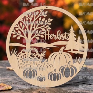 May include: A circular light-coloured wooden cutout featuring a laser-cut design of a tree, a basket of apples, and several pumpkins. The word "Herbst" is inscribed in the centre of the design, which depicts a landscape with rolling hills.