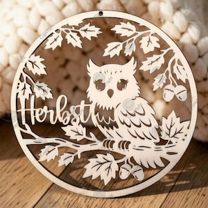May include: A circular light-colored wooden ornament featuring a laser-cut design of an owl perched on a branch. The design includes oak leaves, acorns, and the word "Herbst" in script lettering. A small hole is located at the top of the frame.