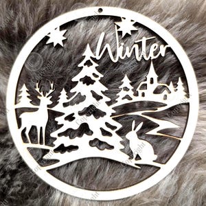 Puede incluir: Adorno de invierno de madera blanca con diseño circular. El adorno presenta un ciervo, un conejo, árboles, una iglesia y la palabra "Winter". También se incluyen estrellas. El adorno tiene un pequeño agujero en la parte superior para colgar.