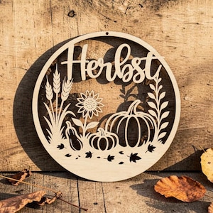 Op de afbeelding: Een rond bord van lichtgekleurd uitgesneden hout met het woord "Herbst" in cursief schrift. Het ontwerp bevat lasergesneden silhouetten van tarwearen, een zonnebloem, twee pompoenen en verspreide bladeren binnen de cirkelvormige lijst.