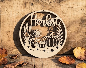 May include: A circular light-colored wooden cutout sign featuring the word "Herbst" in a cursive script. The design includes laser-cut silhouettes of wheat stalks, a sunflower, two pumpkins, and scattered leaves arranged within the circular frame.
