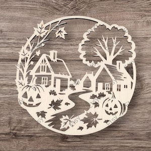 May include: A circular wooden laser-cut artwork depicting an autumnal scene. It shows two houses, a path, trees, falling leaves, and jack-o'-lanterns. The piece is unpainted, displaying the natural colour of the wood.