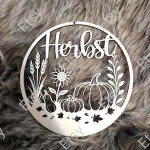Könnte beinhalten: Ein kreisförmiges Metallschild mit dem Wort "Herbst" in Schreibschrift. Das Schild zeigt ausgeschnittene Designs von Weizen, einer Sonnenblume, Kürbissen und Blättern, alles im Herbstthema.