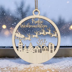May include: A circular light-colored wooden ornament featuring a laser-cut design of Santa Claus in a sleigh pulled by reindeer flying over a village silhouette. The text Frohe Weihnachten is engraved at the top, and it hangs by a twine loop.