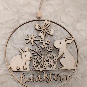 K&ouml;nnte beinhalten: Eine h&ouml;lzerne Osterdekoration mit zwei Hasen, Blumen und Eiern. Die Dekoration ist kreisf&ouml;rmig und tr&auml;gt den Text "Frohe Ostern".