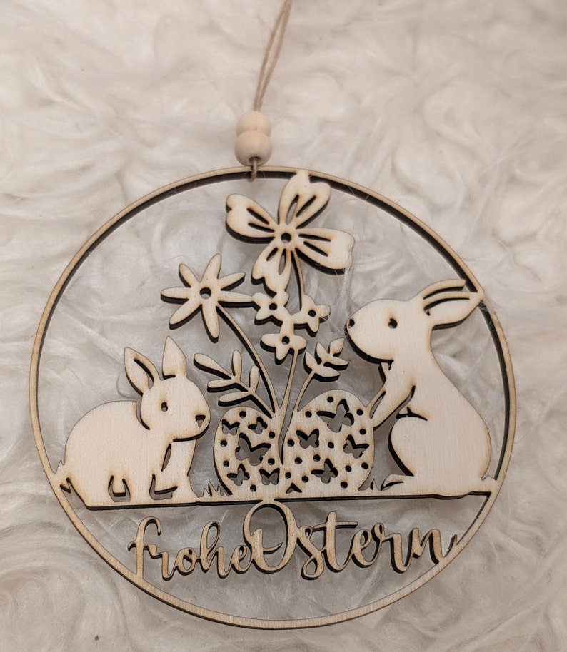 K&ouml;nnte beinhalten: Eine h&ouml;lzerne Osterdekoration mit zwei Hasen, Blumen und Eiern. Die Dekoration ist kreisf&ouml;rmig und tr&auml;gt den Text "Frohe Ostern".