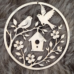 Könnte beinhalten: Runder Holzschmuck mit einem Vogelhaus, zwei Vögeln und floralen Elementen. Das Design ist aus hellem Holz geschnitten, mit einem fliegenden Vogel. Geeignet für die Heimdekoration, hat das Ornament eine natürliche, rustikale Ausstrahlung.