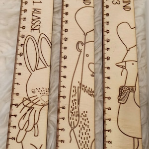 May include: Three wooden rulers with illustrations of a bunny, a bear, and a goose. The rulers are marked with numbers from 1 to 20. The rulers have the text "Du bist 1. Klasse", "Schulkind 2023", and "Schulkind 2023" printed on them.