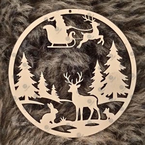 Puede incluir: Un adorno circular de madera clara con un diseño cortado a láser de una escena de bosque invernal. El recorte muestra un trineo con una figura, un reno, pinos, un ciervo de pie, un conejo y un pequeño animal en la base.