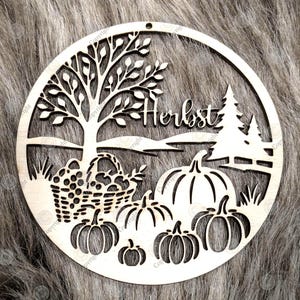 Puede incluir: Decoración otoñal de madera cortada con láser. El diseño presenta un árbol, calabazas, una cesta de frutas y la palabra "Herbst". El adorno circular es de madera clara.