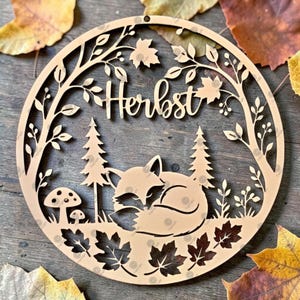 May include: A circular light-colored wooden cutout featuring a laser-cut design of a sleeping fox, two pine trees, two mushrooms, and maple leaves. The word "Herbst" is centered in the upper portion of the circle, framed by branches and foliage.