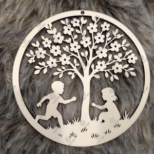 May include: A circular, laser-cut wooden ornament depicting a tree with flowers, two children, and grass. The children are playing under the tree. The ornament has a small hole at the top for hanging.