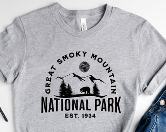 Puede incluir: Camiseta gris con un diseño gráfico negro que dice "Great Smoky Mountain National Park Est. 1934". La camiseta se combina con jeans azules y zapatillas blancas.