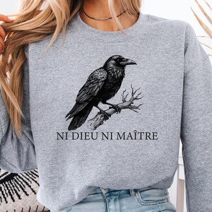 May include: A light gray sweatshirt featuring a detailed black raven perched on a branch. The text "NI DIEU NI MAÎTRE" is printed below the raven. The sweatshirt is a casual style.