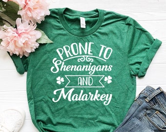 Puede incluir: Camiseta verde con texto blanco que dice "Prone to Shenanigans and Malarkey". La camiseta está doblada y colocada sobre una superficie de madera blanca con un par de zapatillas blancas y unos vaqueros azules.