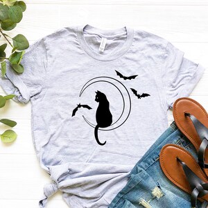 May include: A light gray t-shirt featuring a black cat silhouette sitting on a crescent moon, with three bats flying around it. The shirt is paired with blue jeans and brown sandals.