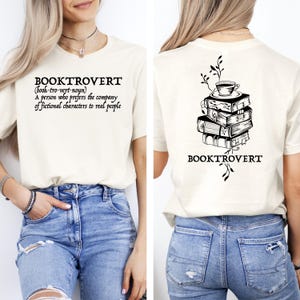 May include: Cream-colored t-shirt with the word "BOOKTROVERT" and its definition printed in black. The back of the shirt features a stack of books with a teacup on top and the word "BOOKTROVERT" below.