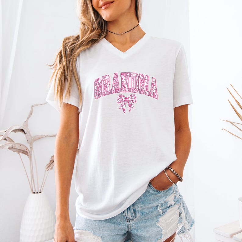 An olive green sweatshirt with "GRANDMA" printed in bold pink floral letters, accented by a matching pink bow. Worn by a woman styled with ripped denim jeans in a cozy, modern setting. A perfect gift for fashionable grandmothers.