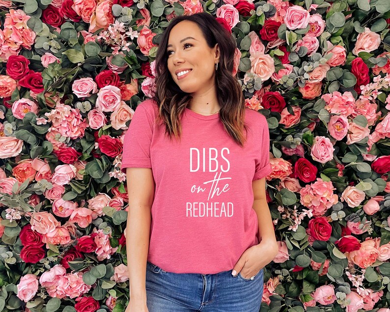 May include: A pink t-shirt with the text "DIBS on the REDHEAD" in white. The person is smiling in front of a wall of pink and red roses and greenery. The shirt is a crew neck style.